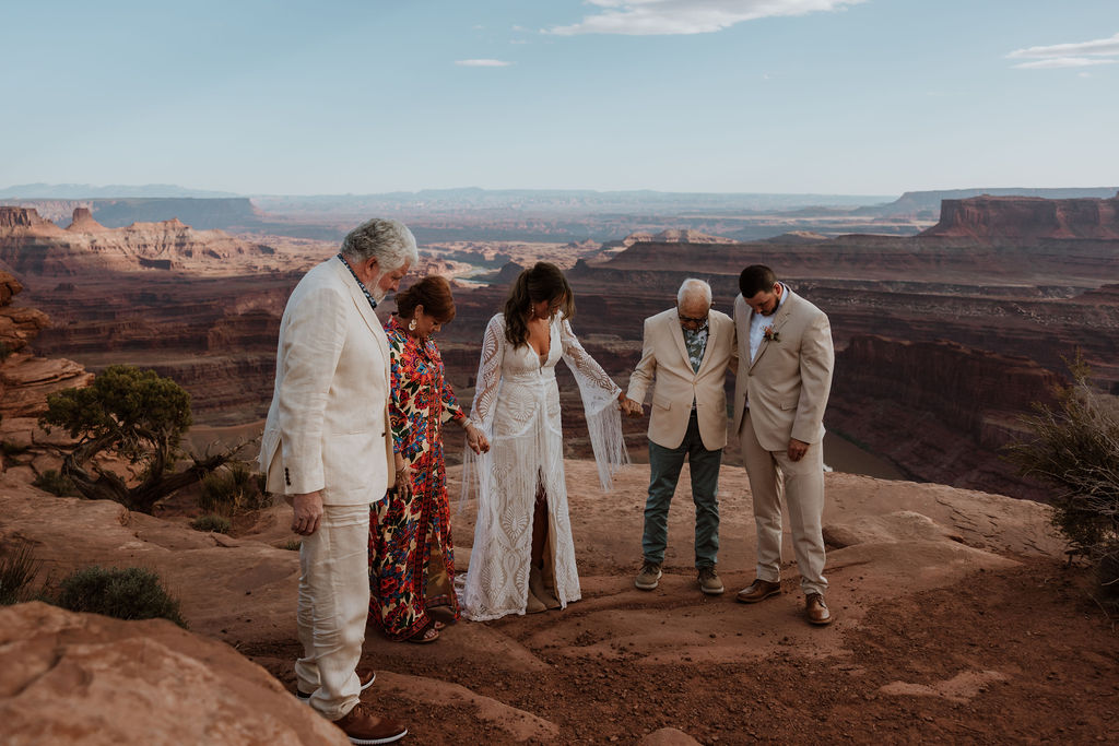 couple holds hands with parents praying at wedding 