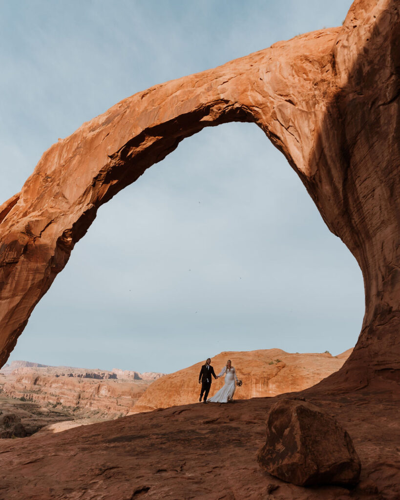 couple walks under arch at outdoor Moab desert elopement