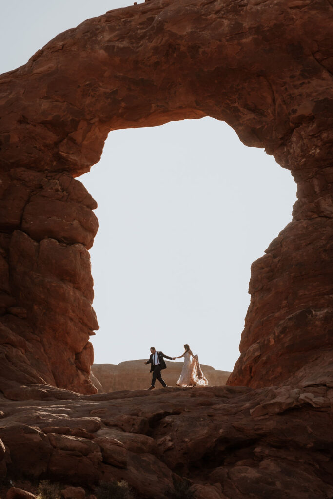 couple walks under arch at outdoor Moab desert elopement