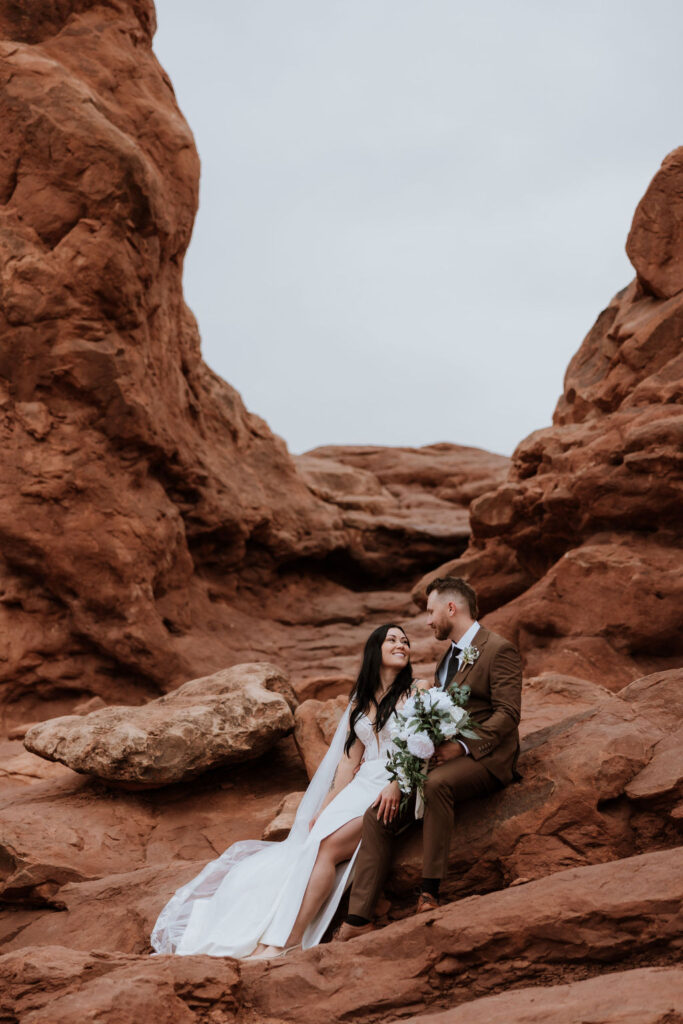 couple sits under arch at Arches National Park elopement