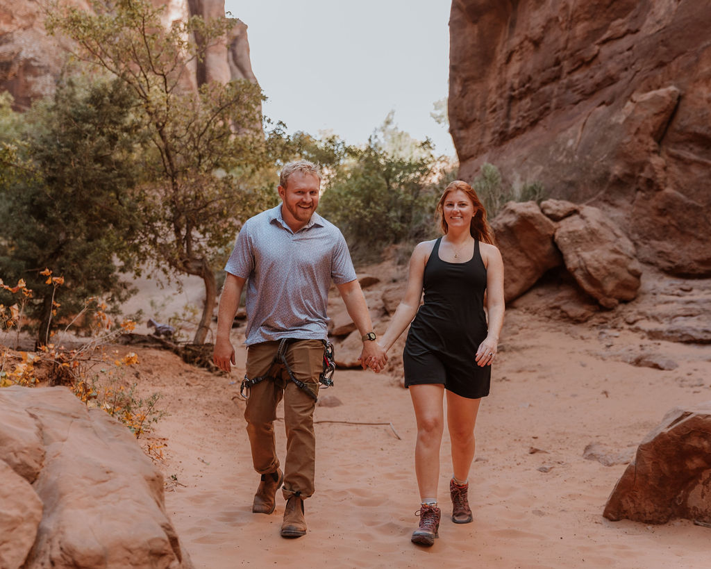 couple walk together holding hands along sandy path in Moab