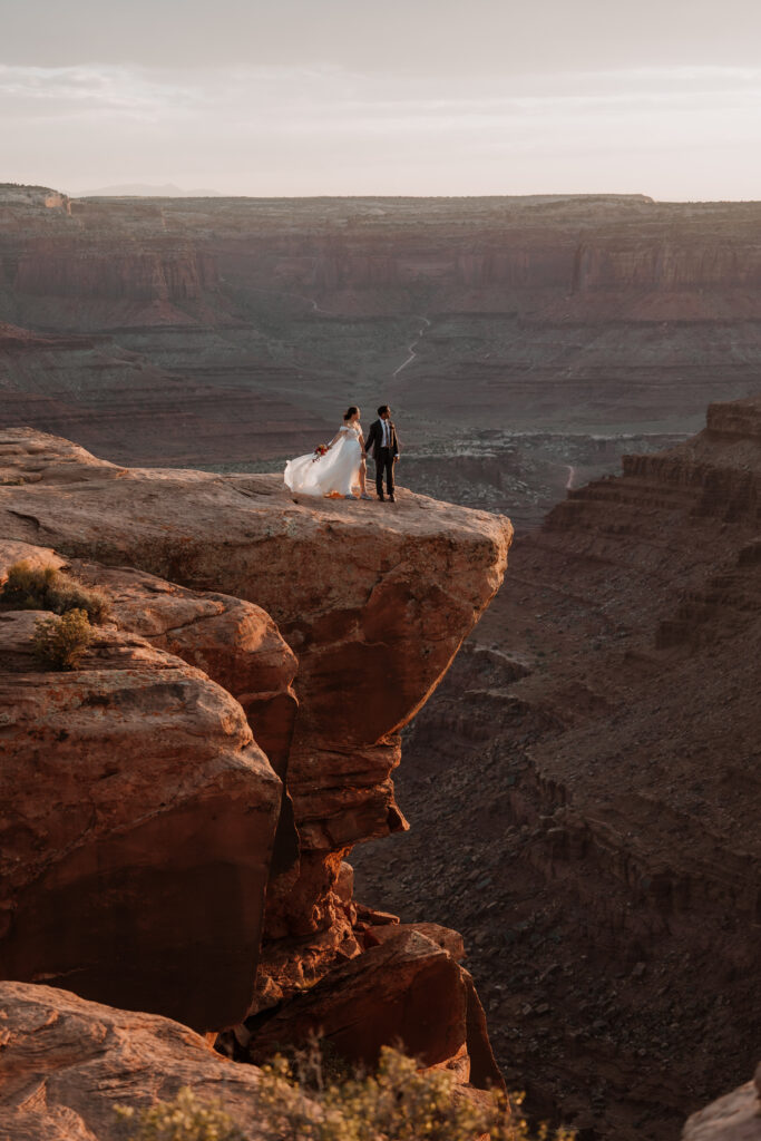 couple stands along rocky ledge of Moab during sunset