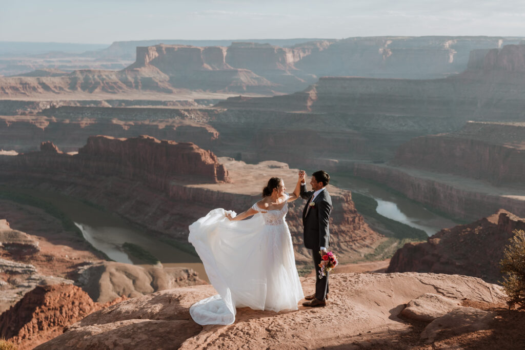 couple hold hands while standing on rocky ledge overlooking Moab's landscape